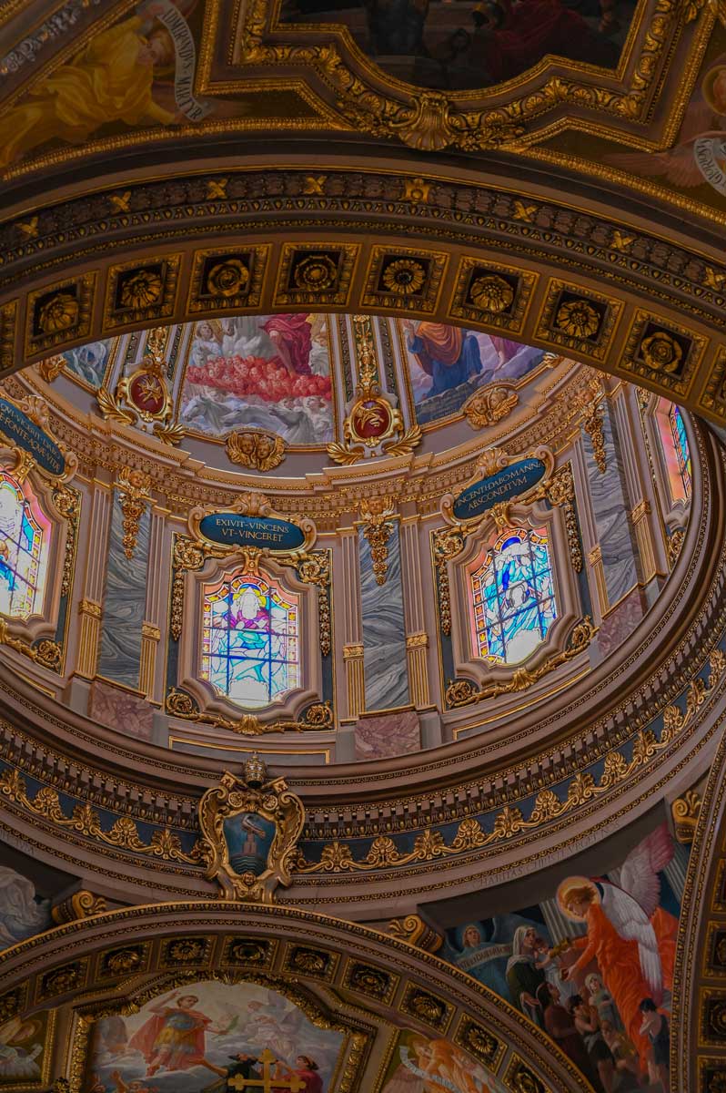 The richly decorated dome interior of St. George’s Basilica in Victoria, Gozo, featuring gold stucco, stained glass, and Baroque frescoes.